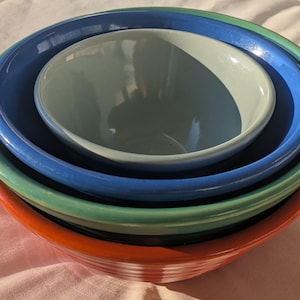 May include: A stack of nested ceramic bowls in various colors: red, green, blue, and a light blue. The bowls are arranged from largest to smallest, creating a visually appealing arrangement. The bowls have a smooth, glossy finish.