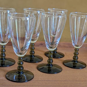 May include: Set of six clear glass wine glasses. Each glass has a flared bowl with vertical ridges, a dark green beaded stem, and a dark green circular base. The glasses are arranged on a wooden surface.