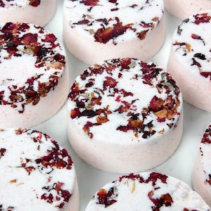 May include: A collection of round, pink bath bombs. Each bath bomb is topped with white granules and scattered with dried red rose petals. The bath bombs are arranged closely together, creating a visually appealing display.