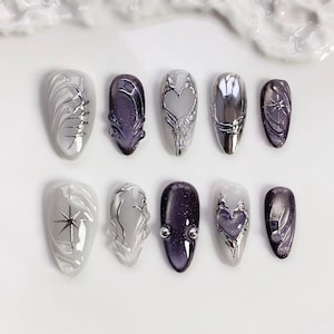 Puede incluir: Un conjunto de diez uñas artificiales con varios diseños. Las uñas presentan una gama de colores, incluyendo plata, gris y morado. Algunas uñas tienen intrincados adornos plateados, mientras que otras muestran diseños en forma de estrella o corazón. Las uñas están dispuestas sobre una superficie blanca.