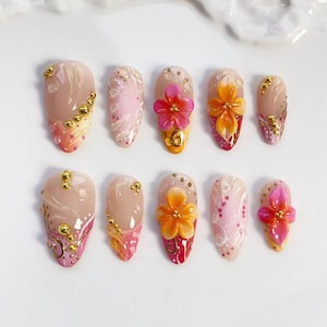 May include: A set of ten artificial nails in various designs. The nails feature a nude base with pink, orange, and gold accents. Some nails have 3D flower embellishments, while others have gold bead details.