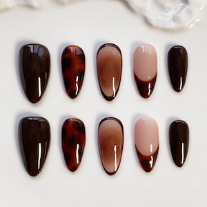 May include: A set of ten artificial nails in different designs and colours. The nails are oval-shaped and feature solid dark brown, tortoiseshell, gradient brown, and a nude with a dark brown French tip. The nails are arranged on a white surface.