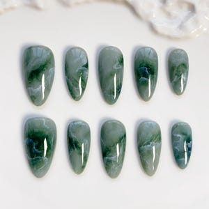 May include: Ten oval-shaped, press-on nails in varying sizes. The nails are a marbled green color with darker green and white swirls, resembling jade. The glossy finish reflects light, enhancing the natural stone appearance. The nails are arranged on a white surface.