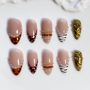 Puede incluir: Un conjunto de diez uñas artificiales con diversos diseños. Las uñas presentan una base nude con detalles como ribetes de perlas, puntas de carey, puntas rojo oscuro, bandas doradas, rayas de cebra y oro texturizado. Dispuestas sobre una superficie blanca.