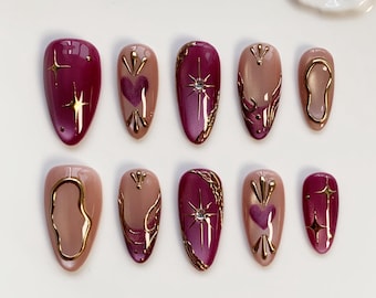 Può includere: Un set di dieci unghie finte a forma di mandorla. Le unghie presentano un mix di colori bordeaux e nude, con dettagli dorati. Alcune unghie hanno disegni a cuore e stella, altre contorni dorati.