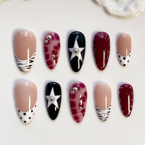 May include: A set of ten artificial nails in various designs. The nails feature a mix of colors, including nude, black, and burgundy, with patterns like zebra stripes, polka dots, and star accents. Some nails are adorned with silver embellishments.