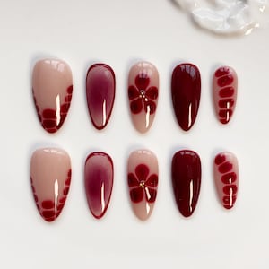 Pode incluir: Um conjunto de dez unhas postiças em forma de amêndoa. As unhas apresentam uma variedade de designs, incluindo bordô sólido, um padrão floral com um centro dourado e um efeito gradiente. A cor de base é um rosa nude.