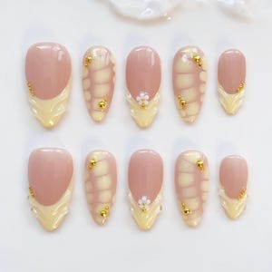 Könnte beinhalten: Ein Set aus zehn künstlichen Nägeln in verschiedenen Größen. Jeder Nagel hat eine hellrosa Basis mit einer hellgelben French-Spitze. Einige Nägel haben ein strukturiertes Design, kleine goldene Perlen und winzige weiße Blumenakzente.