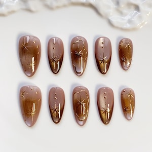 May include: A set of ten almond-shaped false nails. Each nail features a gradient effect, transitioning from a light pink base to a bronze-gold tip. Gold starburst designs adorn each nail, creating a celestial theme.