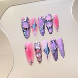 Puede incluir: Conjunto de diez uñas postizas con un diseño degradado en rosa y morado. Cada uña presenta un acento plateado, incluyendo una luna creciente, una estrella y una forma geométrica.