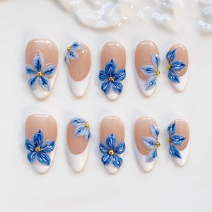 May include: Set of ten artificial nails with a nude base, white tips, and 3D blue flower embellishments. Each flower has a gold center. The nails are oval-shaped and arranged on a white surface.