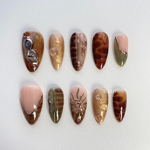 May include: Ten different nail designs featuring various patterns and colours, including brown, gold, green, and white. The designs include a sun, seashells, and abstract patterns.