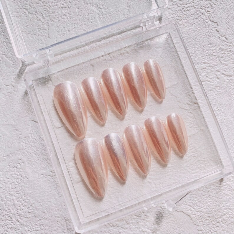 May include: A set of ten pearly white, almond-shaped false nails in a clear plastic case. The nails are arranged in two rows of five.