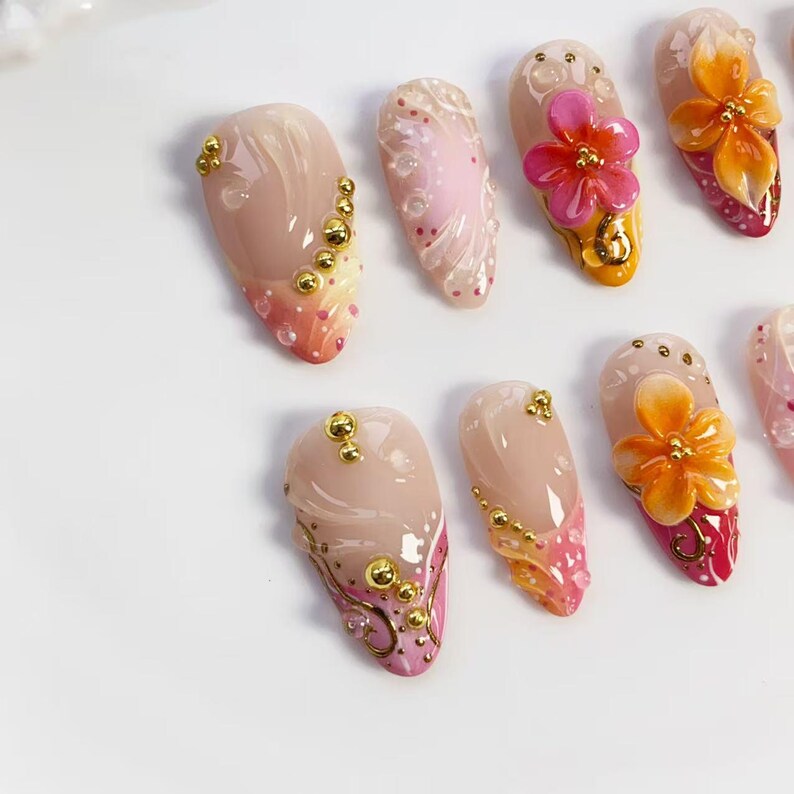 May include: A collection of artificial nails with various designs. Some nails feature gold embellishments, while others showcase floral accents in shades of pink and orange. The nails have a glossy finish and are arranged on a white surface.