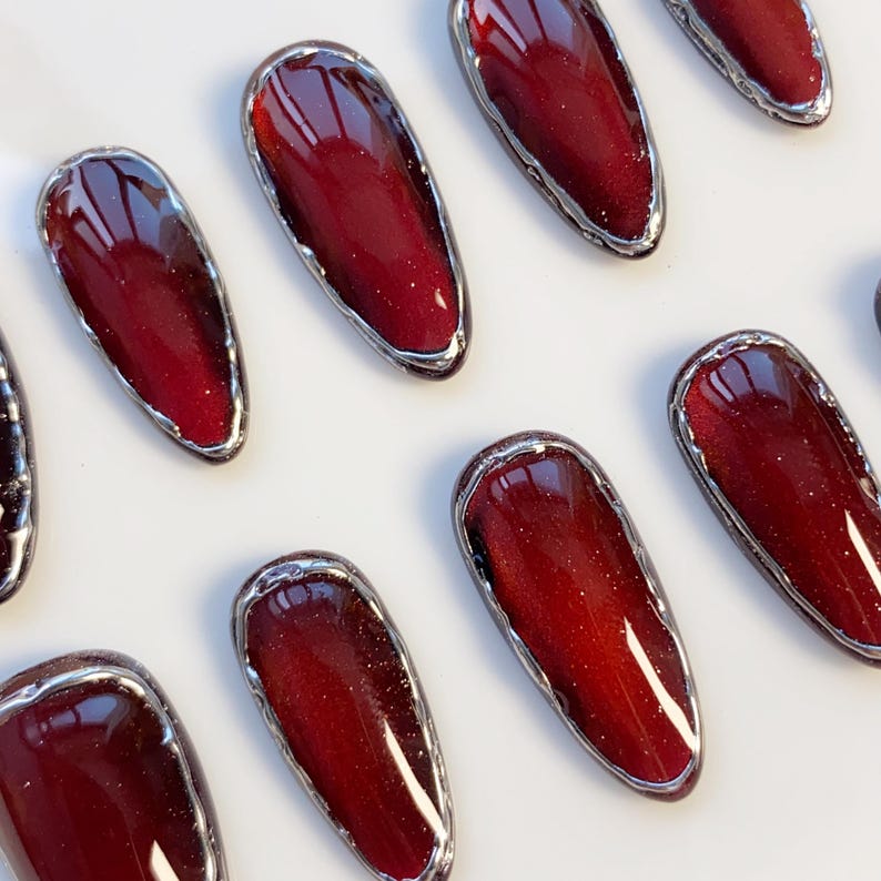 May include: Close-up of several oval-shaped, burgundy-colored press-on nails with a silver metallic border. The nails have a glossy finish and a subtle glitter effect, arranged on a white surface. The design is elegant and suitable for various occasions.