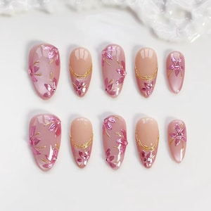 Puede incluir: Un juego de diez uñas artificiales de varios tamaños. Las uñas son principalmente rosas con un acabado brillante. Algunas uñas presentan diseños florales rosas y detalles dorados. Otras tienen una base nude con detalles dorados.
