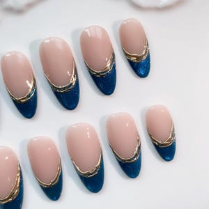 May include: Oval-shaped artificial nails with a light pink base, a gold trim, and a deep blue glitter tip. The nails are arranged on a white surface, showcasing a modern manicure design.