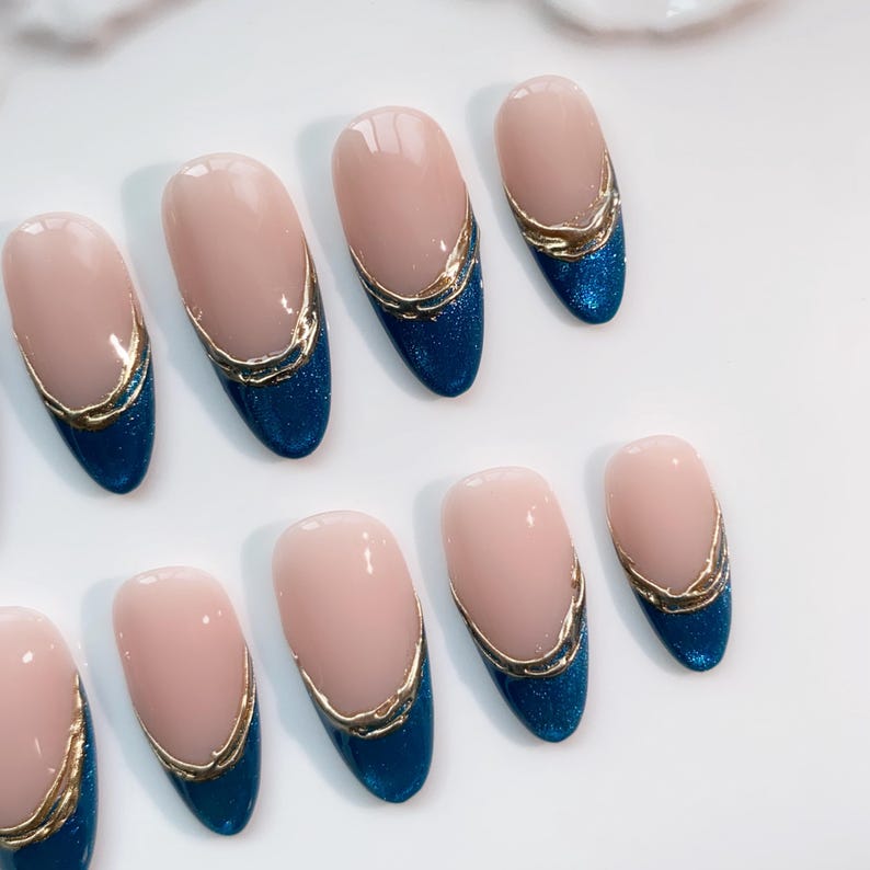 May include: Oval-shaped artificial nails with a light pink base, a gold trim, and a deep blue glitter tip. The nails are arranged on a white surface, showcasing a modern manicure design.