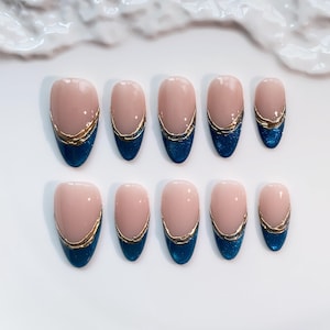 May include: Set of ten oval-shaped false nails. Each nail features a nude base, a gold metallic trim, and a dark blue glittery tip. The nails are arranged on a white surface.