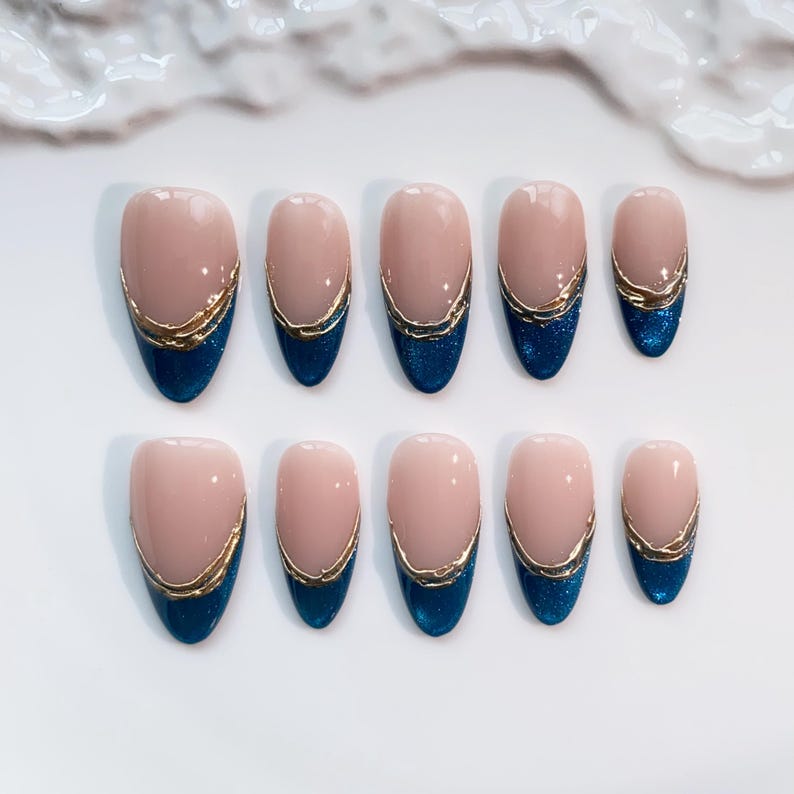 May include: Set of ten oval-shaped false nails. Each nail features a nude base, a gold metallic trim, and a dark blue glittery tip. The nails are arranged on a white surface.