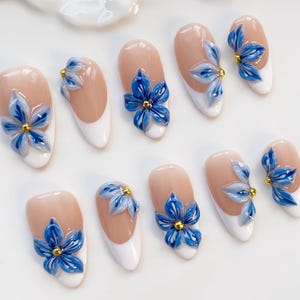 May include: A set of artificial nails with a French manicure design. Each nail features a light pink base, white tips, and a 3D blue flower with a gold center. The flowers have detailed petals and add a touch of elegance.
