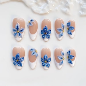 May include: Set of ten artificial nails with a nude base, white tips, and 3D blue flower designs with gold centers. The nails are oval-shaped and arranged on a white surface.