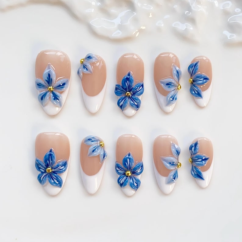 May include: Set of ten artificial nails with a nude base, white tips, and 3D blue flower designs with gold centers. The nails are oval-shaped and arranged on a white surface.