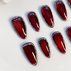May include: A collection of oval-shaped, press-on nails. Each nail features a deep burgundy color with a silver metallic border. The nails are arranged on a white surface, showcasing their glossy finish and design.