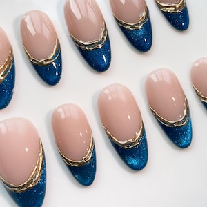 May include: Oval-shaped artificial nails with a light pink base and a deep blue glitter tip. A gold-colored decorative line separates the two colors. The nails are arranged on a white surface.
