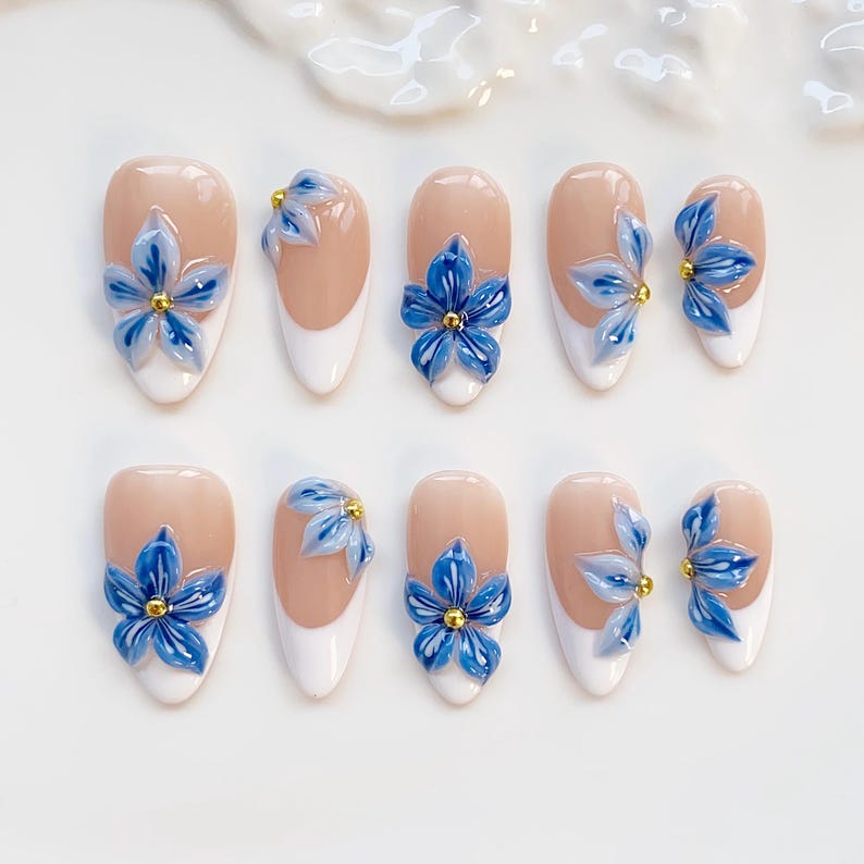 May include: Set of ten artificial nails with a light beige base, white tips, and blue flower embellishments. Each flower has a gold center. The nails are oval-shaped and arranged on a white surface.