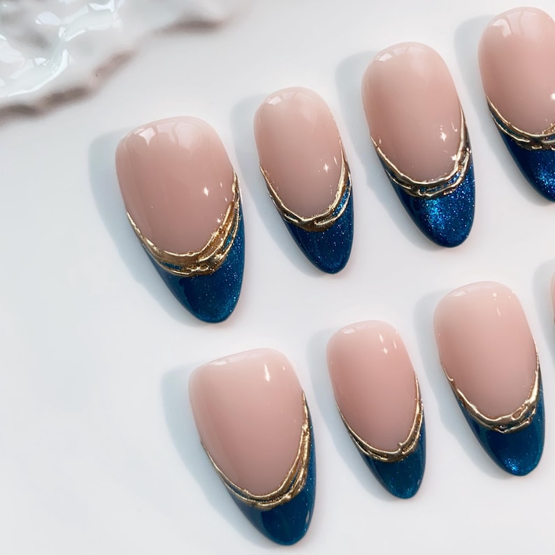 May include: Oval-shaped artificial nails with a light pink base, a gold trim, and a dark blue glitter tip. The nails are arranged on a white surface, showcasing a classic French manicure design.
