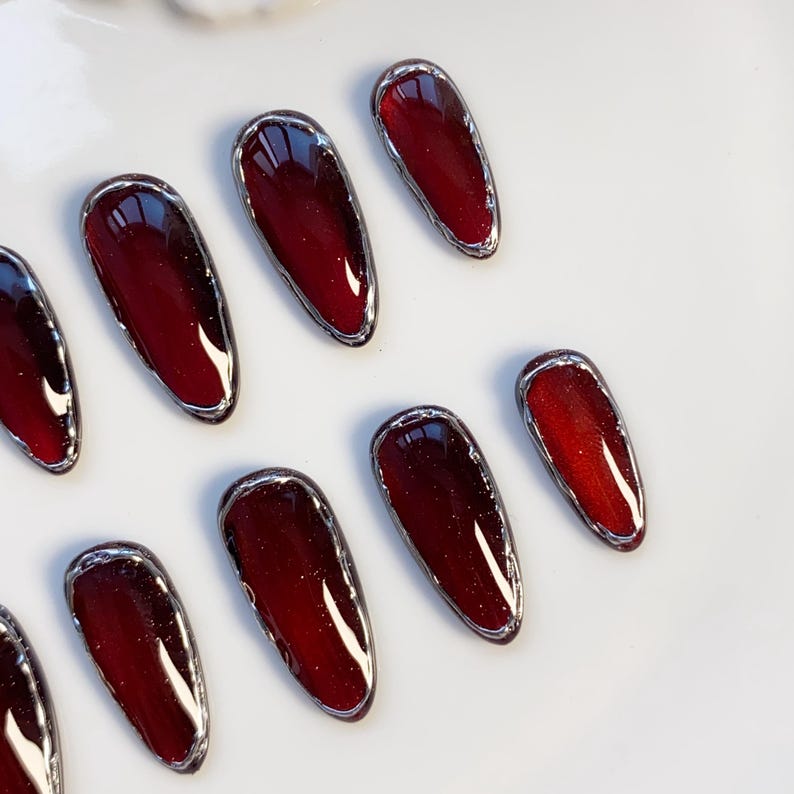 May include: A collection of burgundy-colored, oval-shaped false nails with a silver metallic border. The nails have a glossy finish and are arranged on a white surface. The nail tips are a darker shade of burgundy.