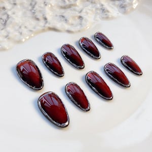 May include: Set of ten burgundy-colored, oval-shaped false nails with a silver metallic trim. The nails are arranged on a white surface, showcasing their glossy finish and elegant design. A beauty product for nail art.