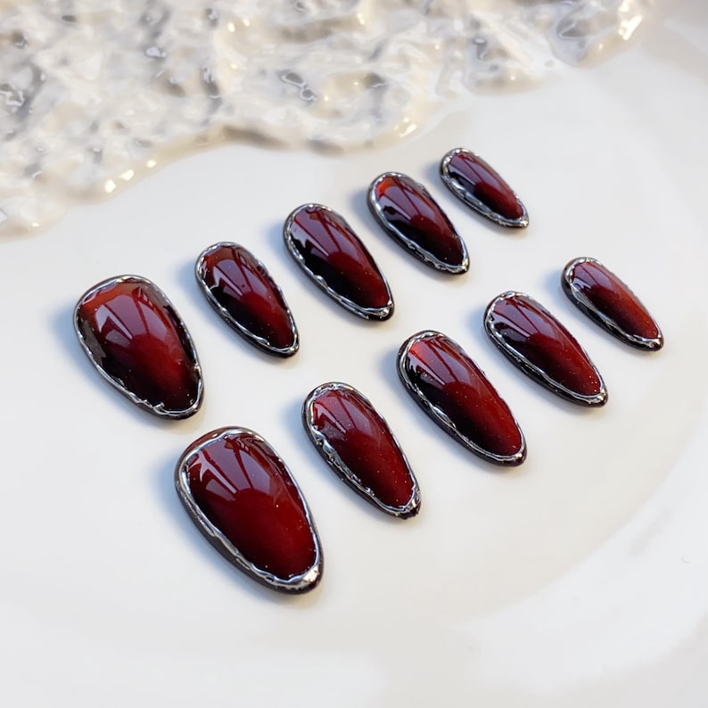 May include: Set of ten burgundy-colored, oval-shaped false nails with a silver metallic trim. The nails are arranged on a white surface, showcasing their glossy finish and elegant design. A beauty product for nail art.