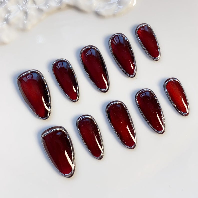 May include: Set of ten burgundy-colored, oval-shaped false nails with a silver metallic trim. The nails have a glossy finish and are arranged on a white surface. The nail set is a beauty product.