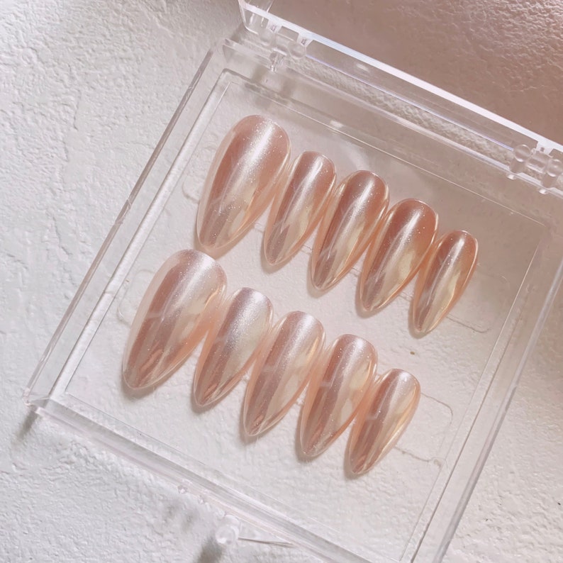 May include: A set of ten nude-colored, long, almond-shaped press-on nails with a pearlescent shimmer finish.