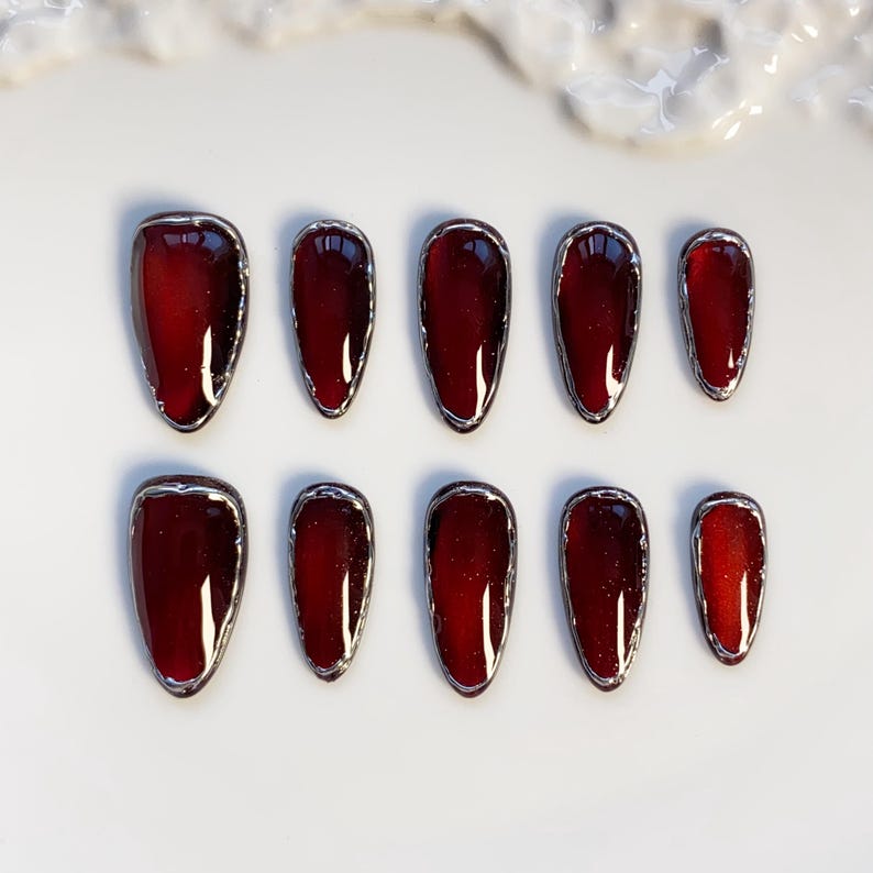 May include: Set of ten burgundy-colored, oval-shaped false nails with a silver metallic trim. The nails are arranged in two rows of five on a white surface, showcasing a glossy finish and a modern aesthetic.
