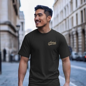 Könnte beinhalten: Schwarzes T-Shirt mit Rundhalsausschnitt und goldenem Text "Arid Drall Urchin U6OT". Das Shirt besteht aus einem weichen Material und eignet sich für Freizeitkleidung.