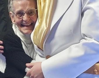 May include: A man in a white suit with a gold scarf is embraced by an older woman. The man's suit has a wide collar and gold buttons. The woman wears glasses and a black and white striped sweater.