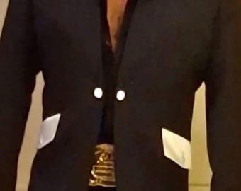 May include: A black blazer featuring two white buttons at the center closure and two white pocket flaps. The blazer is worn over a black shirt and black trousers, accented by a wide, metallic gold belt with a prominent, ornate rectangular buckle.