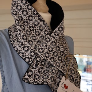 May include: A patterned cowl scarf with a black and white geometric design. The scarf is displayed on a mannequin, showcasing its shape and style. The tag reads "fabs Handmade with love."