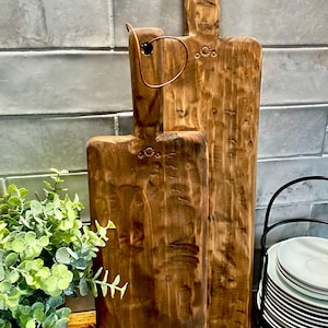 May include: Two rustic wooden cutting boards with a brown finish and a copper wire handle. The boards are hanging on a white tiled wall.