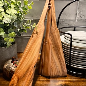 May include: Three wooden kitchen spatulas with a natural wood grain finish. The spatulas are hanging together by a brown leather loop. The spatulas have a rounded, paddle-shaped head.