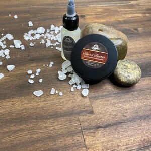 May include: A collection of beard care products, including a bottle of beard oil, a tin of beard butter, and several small, smooth stones. The beard butter tin has a label that reads "Board Butter". The products are arranged on a wooden surface.
