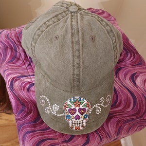 May include: A green canvas baseball cap with a white and colorful sugar skull design. The skull has rhinestones around it.