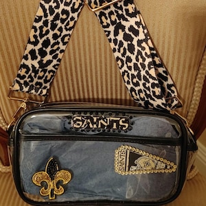 May include: Clear plastic crossbody bag with a black and white leopard print strap. The bag has a blue denim lining with gold and black embellishments and the word "Saints" in rhinestones.