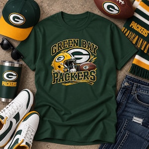 Puede incluir: Una camiseta verde oscuro con un gráfico de un casco de fútbol americano de los Green Bay Packers y un balón de fútbol con el texto "GREEN BAY PACKERS" en letras amarillas y blancas. La camiseta está rodeada por una gorra, gafas de sol, un vaso y una bufanda.