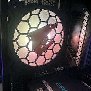 Puede incluir: Primer plano de un ventilador de ordenador con diseño de panal y logotipo rojo. El ventilador está iluminado con un degradado de colores, incluyendo amarillo, rosa y blanco. También se ve una tarjeta gráfica con la palabra "EAGLE" en azul.