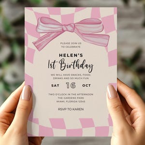 Könnte beinhalten: Eine rosa und weiße Geburtstagseinladung mit einer Schleifengrafik. Auf der Einladung steht "Helens 1. Geburtstag" und enthält Veranstaltungsdetails wie Datum, Uhrzeit und Ort. Die Einladung wird von einer Person gehalten.