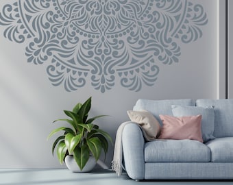 May include: A light blue sofa with two pillows, one white and one pink, sits in front of a wall with a large gray mandala stencil design. A potted green plant is on the left side of the sofa.